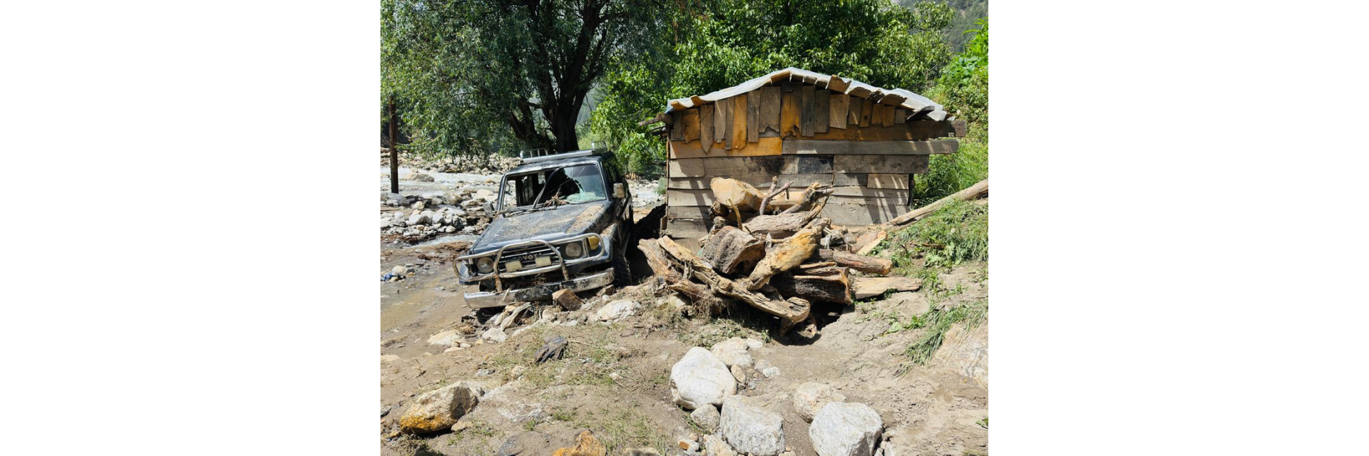 A vehicle covered in mud beside a damaged wooden structure surrounded by debris, rocks and fallen tree trunks after flooding in Afghanistan.