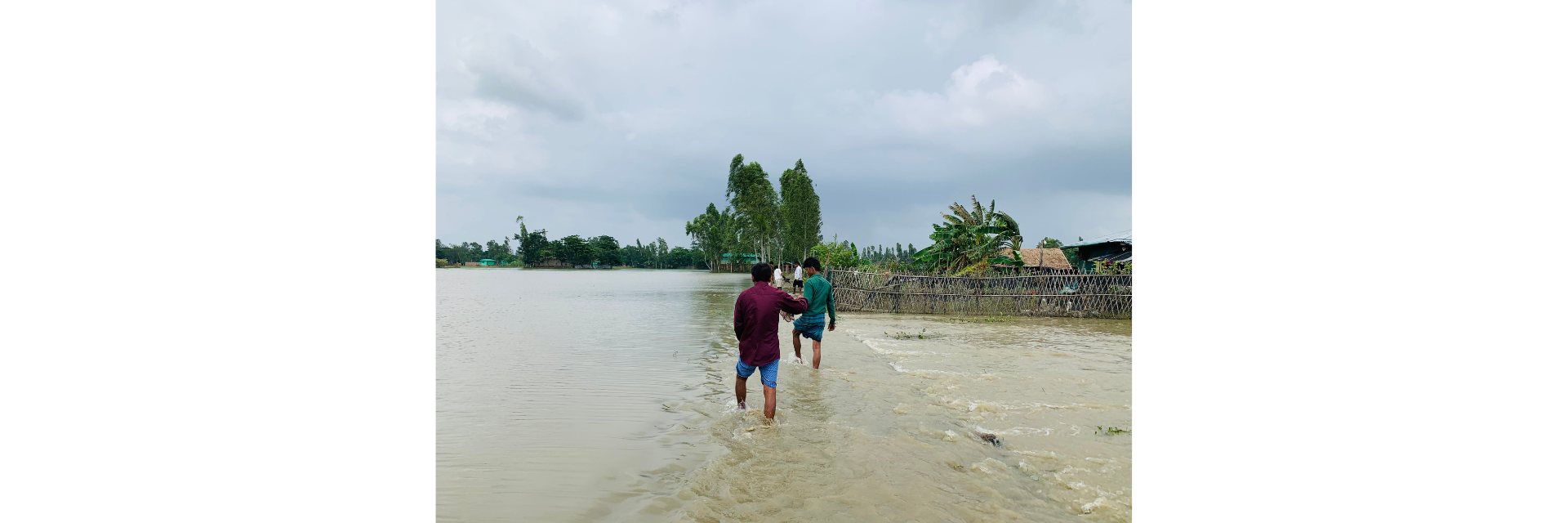 Two people walking through floodwaters in Bangladesh, with flooded homes and trees in the background.