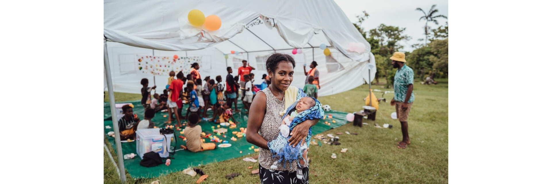 Rael and her baby stand in front of a tent where children are playing