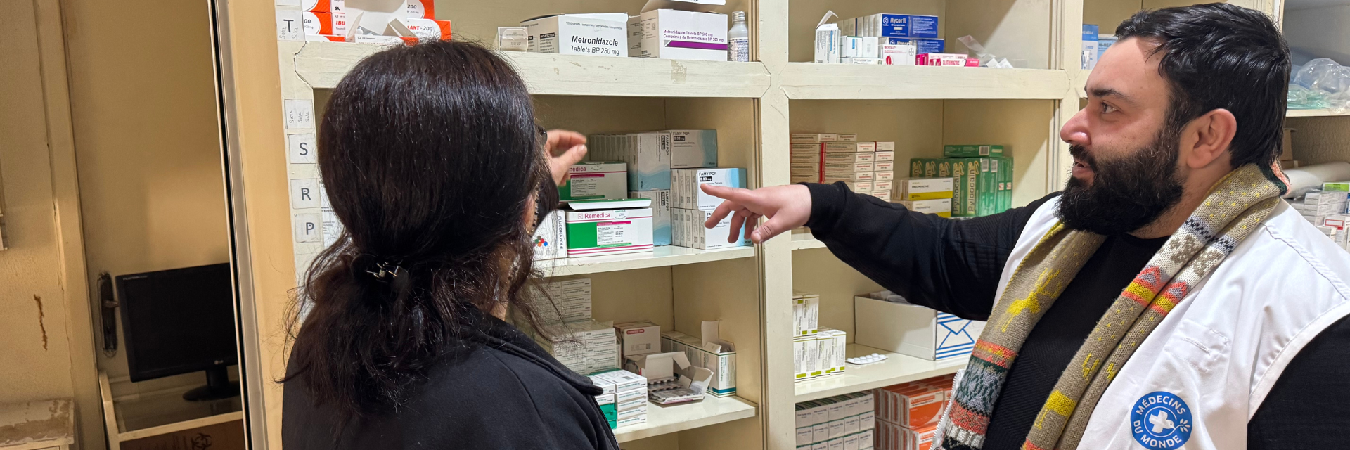 Man wearing a Médecins du Monde vest gestures at shelves of medical supplies while talking to a woman.