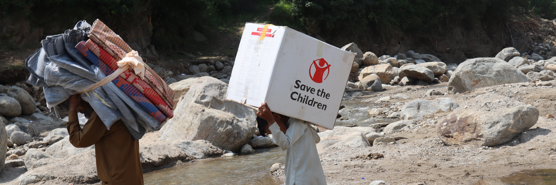 Two men carry supplies across a stream. One carries textiles, another has a box that reads "Save the Children"