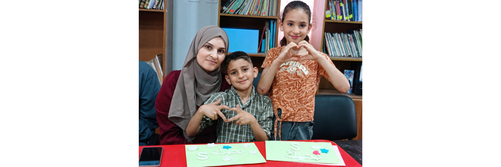 A mother and her two children making hearts with their hands while creating handmade cards at the Makani Club.