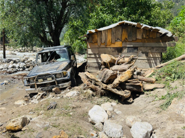 A vehicle covered in mud beside a damaged wooden structure surrounded by debris, rocks and fallen tree trunks after flooding in Afghanistan.