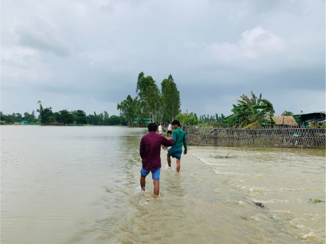 Two people walking through floodwaters in Bangladesh, with flooded homes and trees in the background.