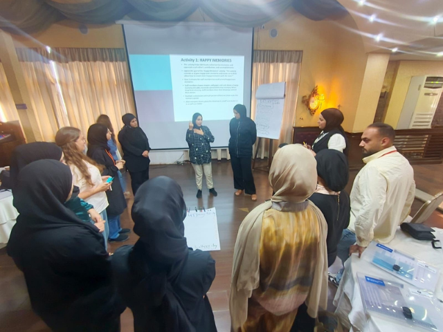 A group of people stand in a circle. A document on a projector reads "Activity one: Happy memories"
